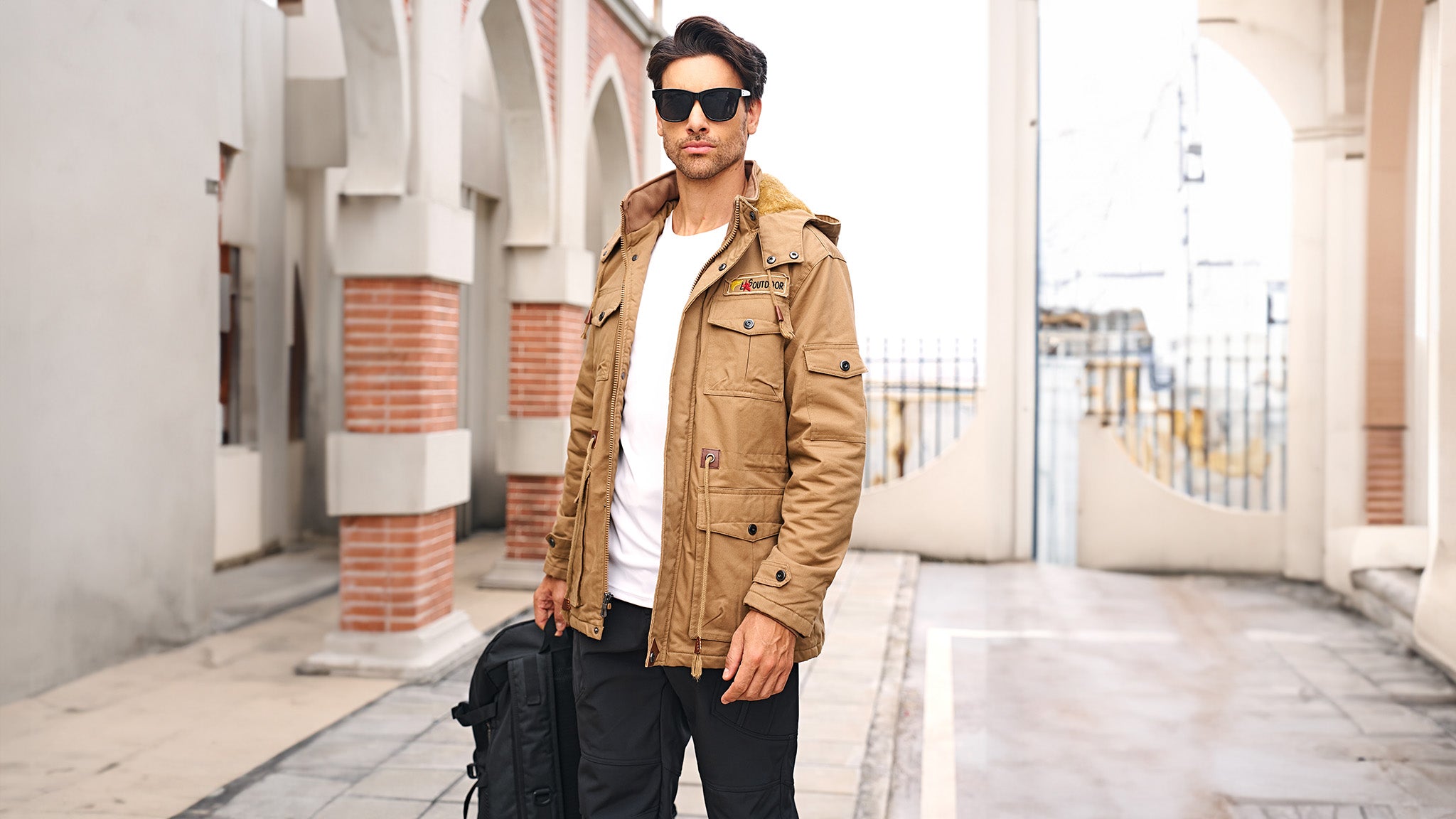 Man in a tan jacket and sunglasses holding a black backpack in an urban setting.